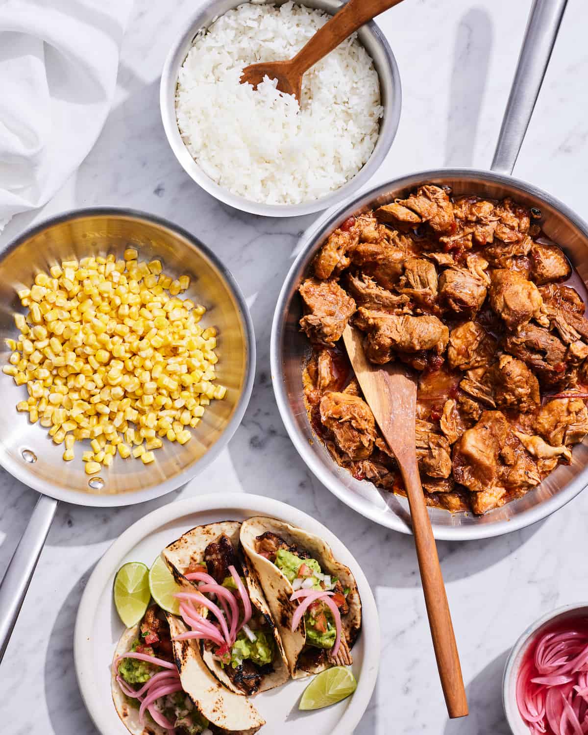 A skillet with carnitas, with a small skillet of corn, a bowl of white rice and a plate of tacos with a small bowl of pickled onions on the side.