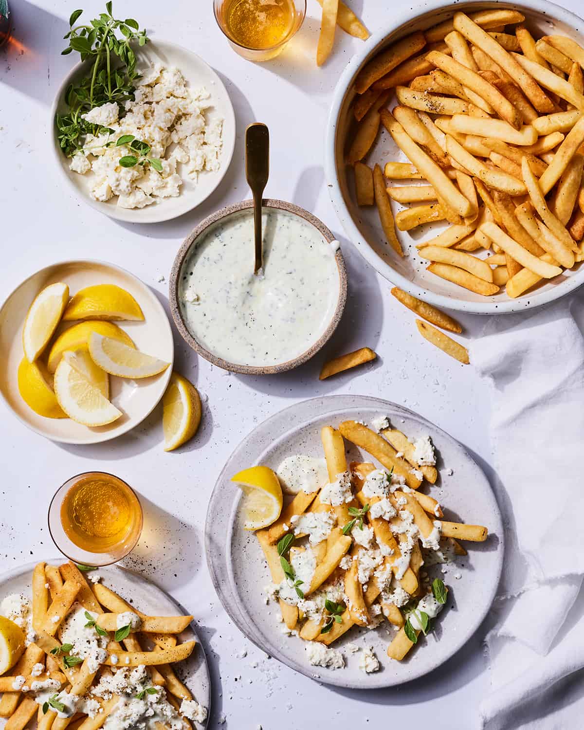 A bowl of homemade fries next to a bowl of feta dipping sauce, a bowl of feta, and a bowl of lemon wedges.  
