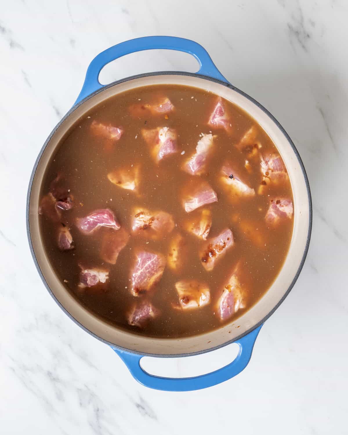 A large blue dutch oven with pork in a mixture of water, seasonings and orange juice.