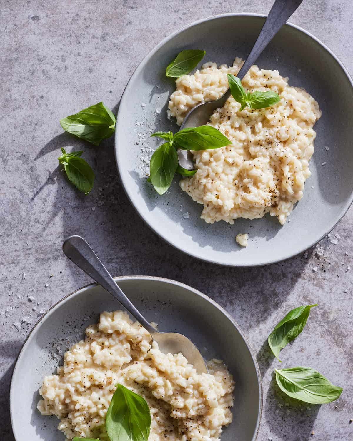an overhead shot of two shallow grey bowls of goat cheese risotto sitting on a grey concrete tabletop. the risotto is garnished with freshly cracked black pepper salt and basil leaves. a silver spoon rests on the edge of the bowl
