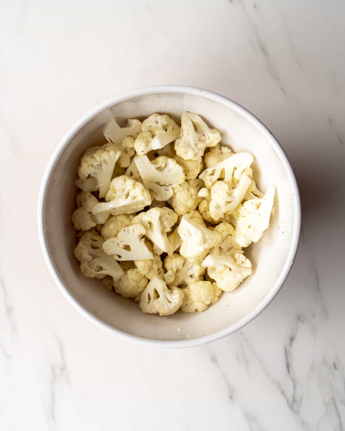 A white bowl with cut cauliflower florets tossed with oil and seasoning.