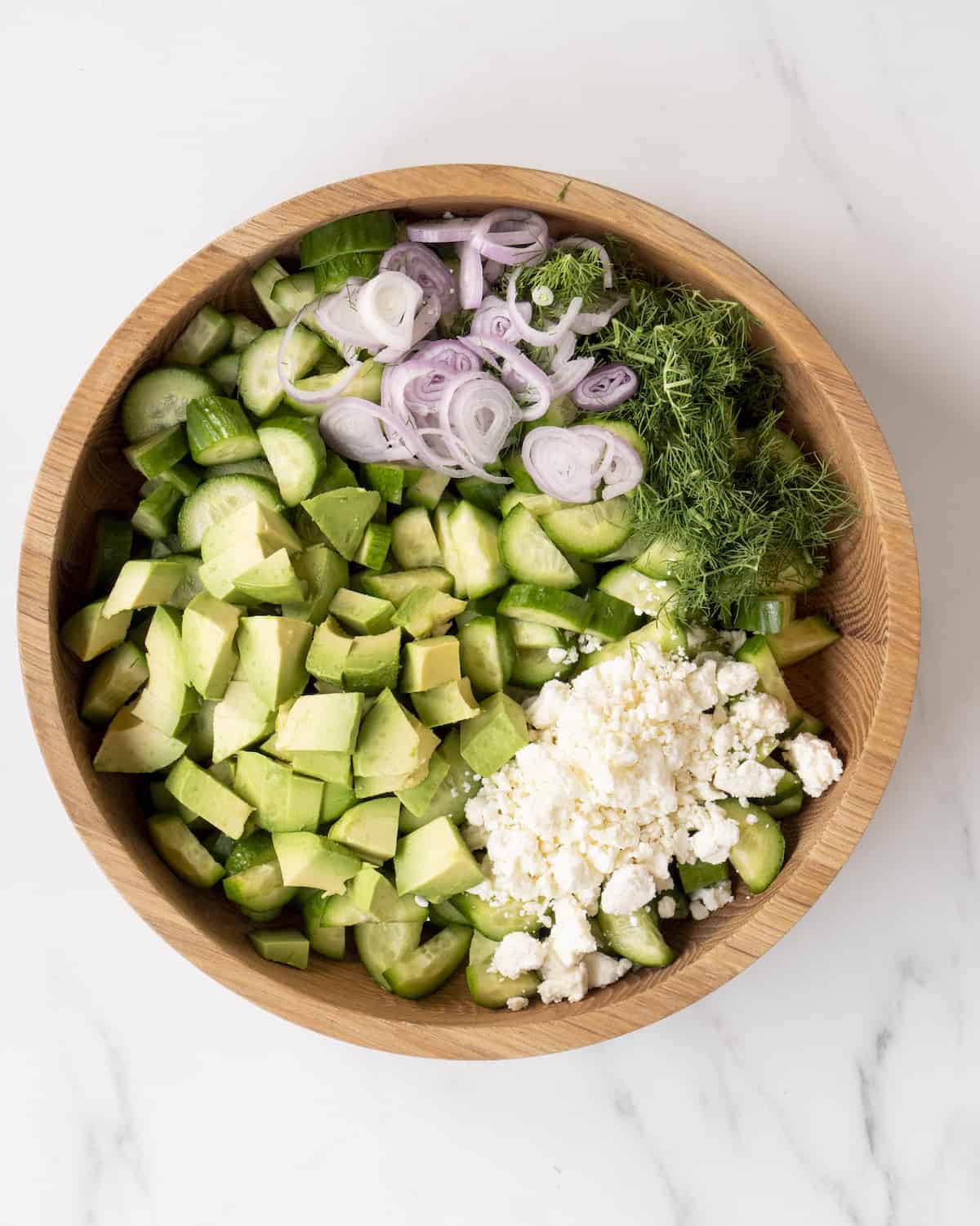 A wooden mixing bowl with chopped up cucumber and avocado, feta cheese, dill and sliced shallots.