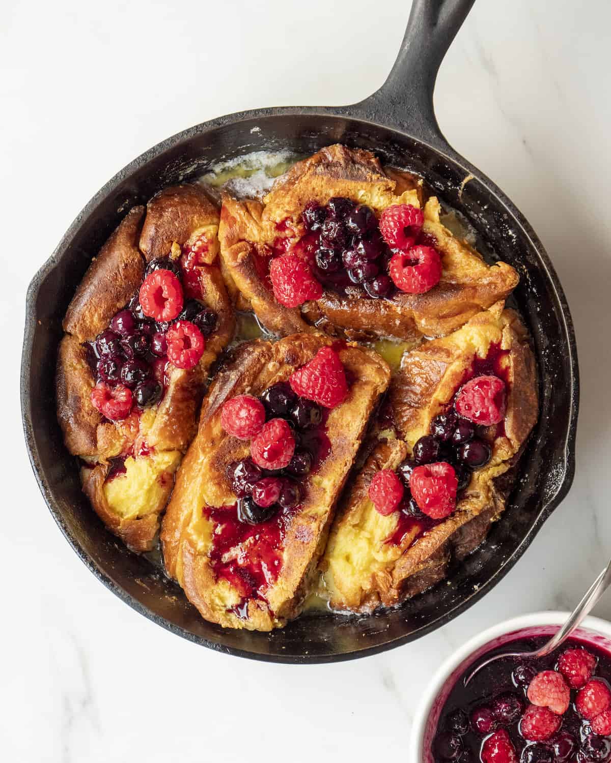 Cooked french toast in a skillet, with berry compote on top and a bowl of berry compote on the side.