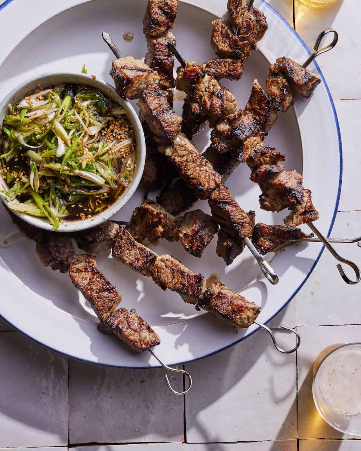 8 grilled steak skewers on a large white circle serving plate and a small bowl of scallion dipping sauce nestled between the skewers on the plate.