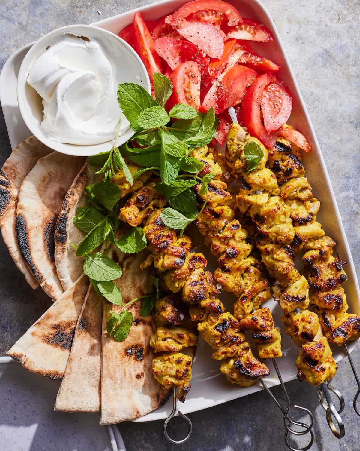 A white tray on a concrete table with 5 moroccan chicken skewers garnished with mint, pita bread, tomato wedges, and small bowl of whipped greek yogurt