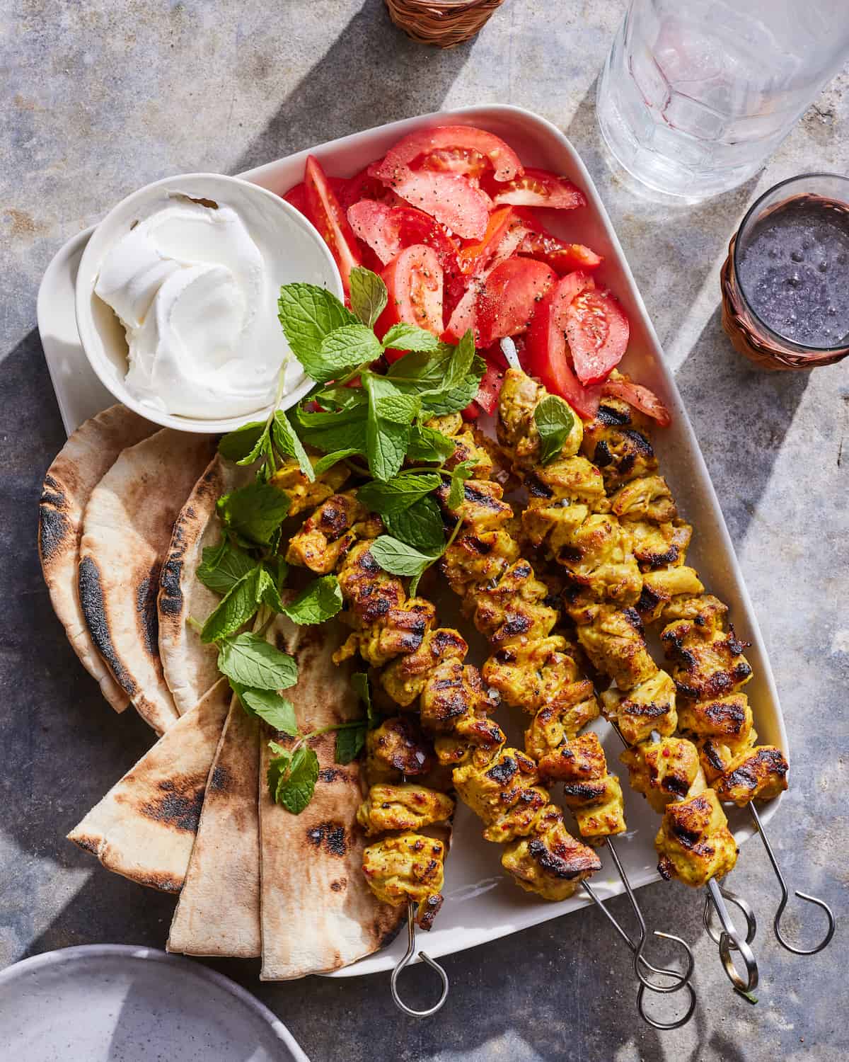 A white tray on a concrete table with 5 moroccan chicken skewers garnished with mint, pita bread, tomato wedges, and small bowl of whipped greek yogurt