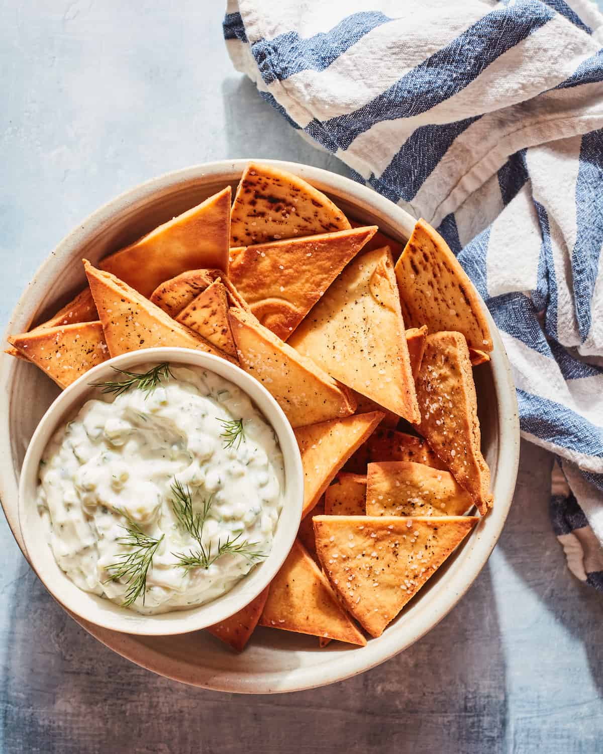 A beige ceramic bowl of triangle pita with a small bowl of tzatziki topped with dill with a white and blue striped kitchen towel.