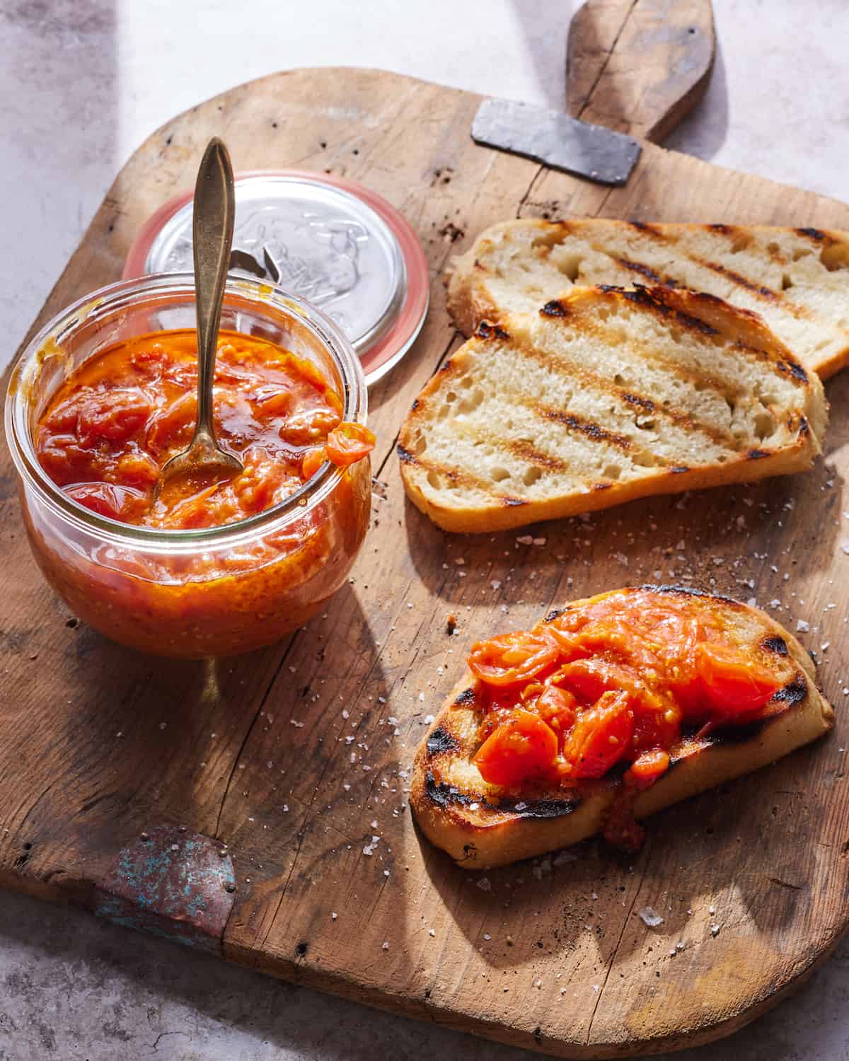 Tomato confit in a mason jar sitting on top of a cutting board next to toast.  