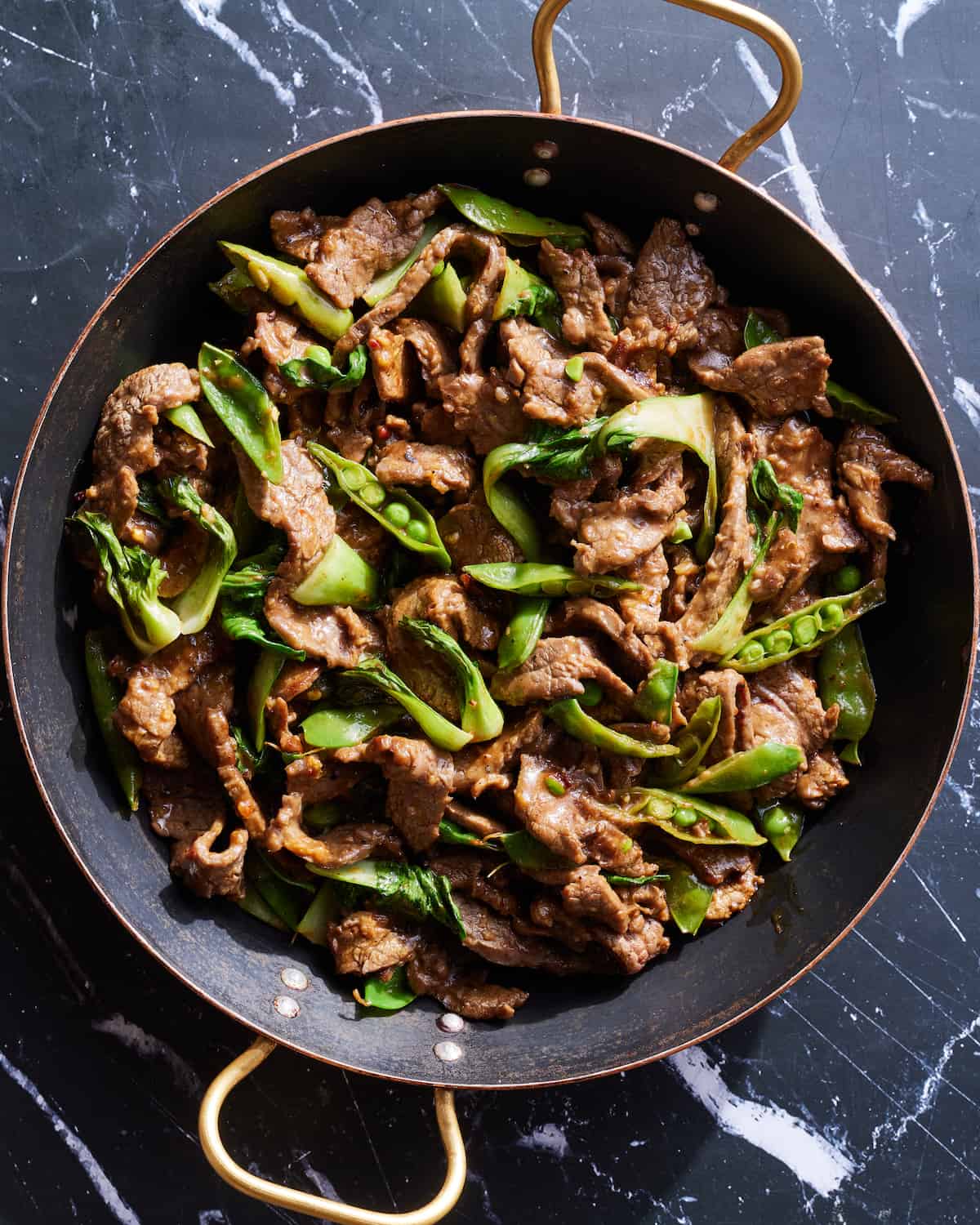 an overhead shot of a large copper braiser of garlic ginger steak stir fry on a black marble table