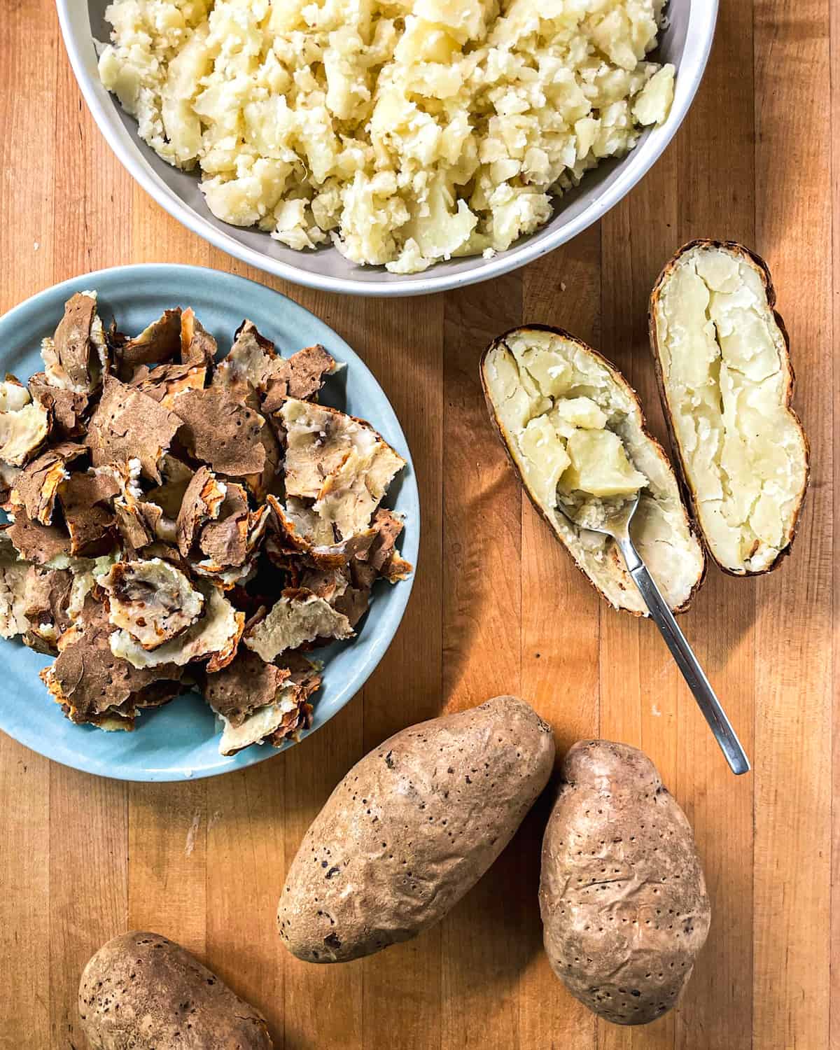 4 Baked russet potatoes with one halved and scooped out next to a large bowl of roughly chopped potato insides and a bowl of potato skins beside.