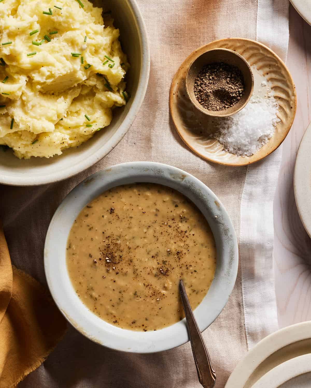 A grey bowl with turkey gravy garnished with freshly ground black pepper with a spoon in it, along with a bowl of mashed potatoes, and some kosher salt and black pepper on the side, all placed on a brown cloth on a wooden table.