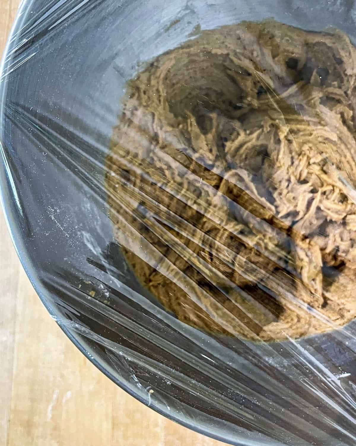 Molasses Cookie dough in a bowl of a stand mixer covered in plastic wrap