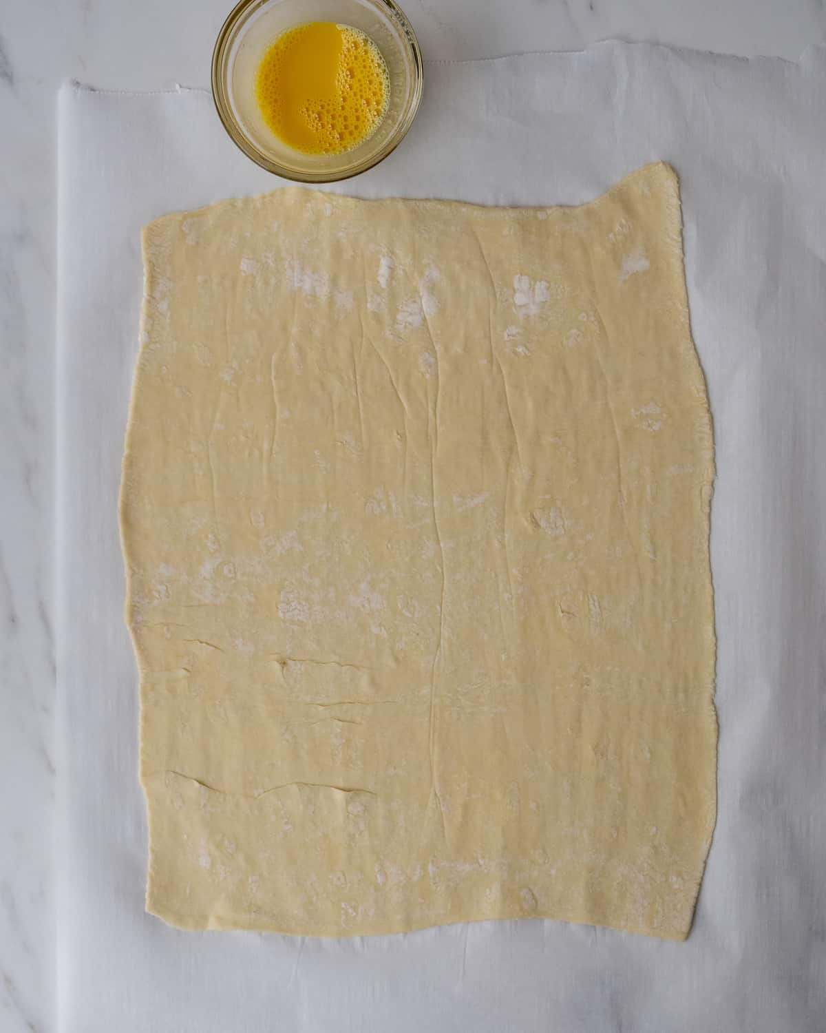 Puff pastry in an 11 x 15-inch rectangle on top of parchment paper with a small clear bowl of beaten egg yolk.