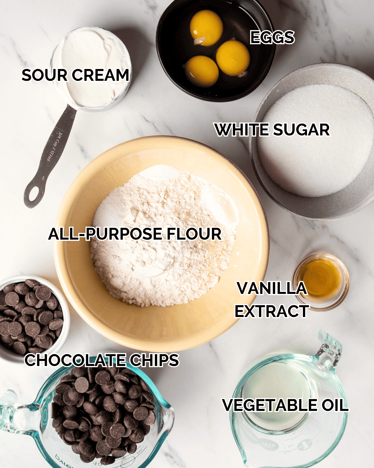 An overhead shot of all the individual ingredients in bowls.  Ingredients displayed in bowls are sour cream, eggs, white sugar, AP flour, chocolate chips, vanilla extract, and vegetable oil.  