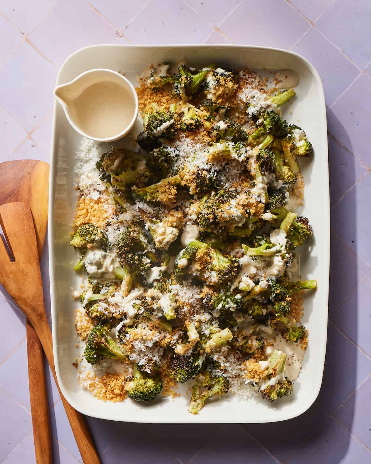 An overhead shot of a white tray of Caesar Roasted Broccoli with a garnish of panko, freshly grated parmesan, and a small bowl of tahini in the top left corner with two wooden serving spatulas laying on the purple tile tabletop.