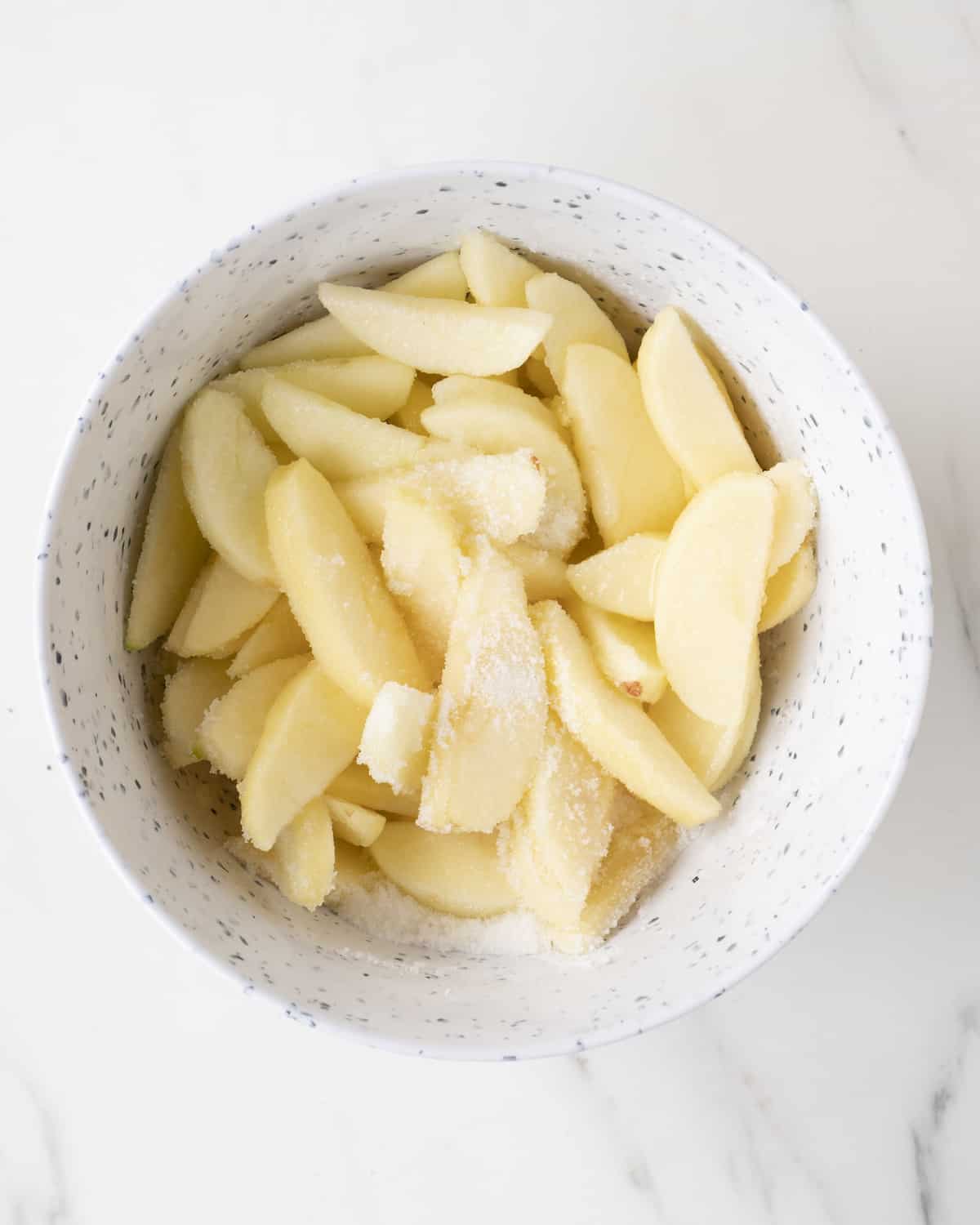 A white mixing bowl with peeled apple wedges, with sugar and lemon juice poured on them.