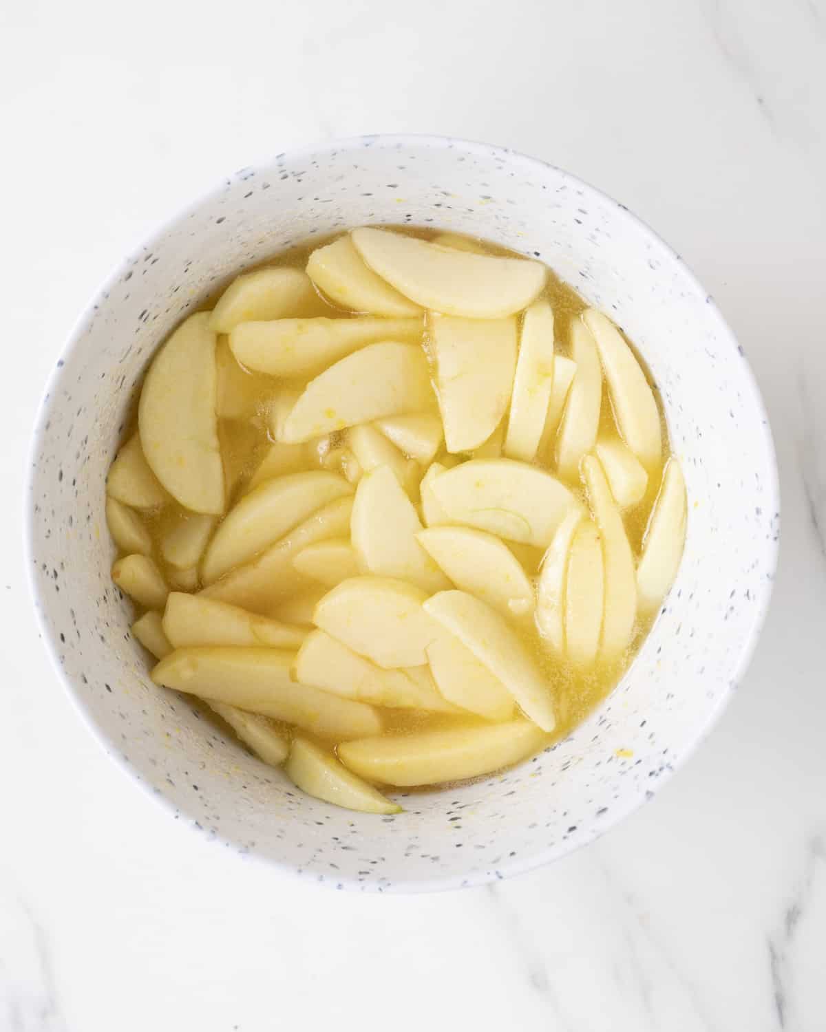 A white mixing bowl with peeled apple wedges in an oil and eggs mixture.