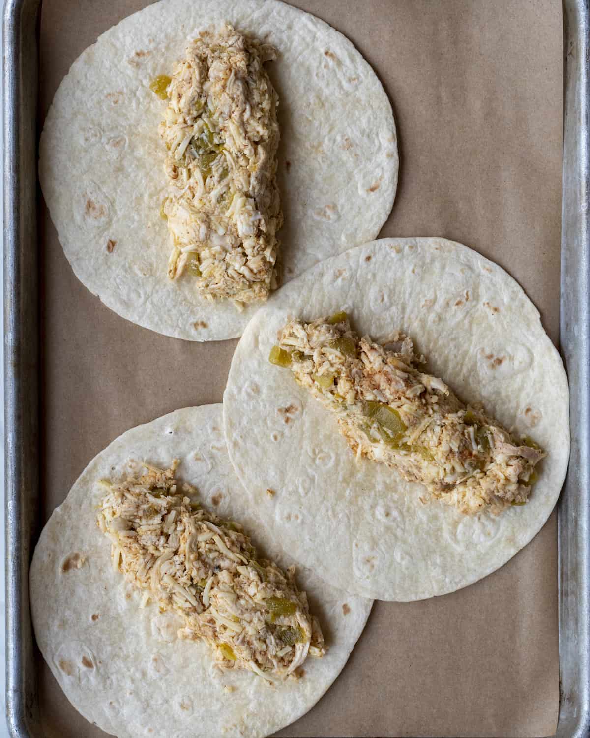 A baking tray lined with parchment paper, with three flour tortillas with the shredded chicken mixture in the center of each.