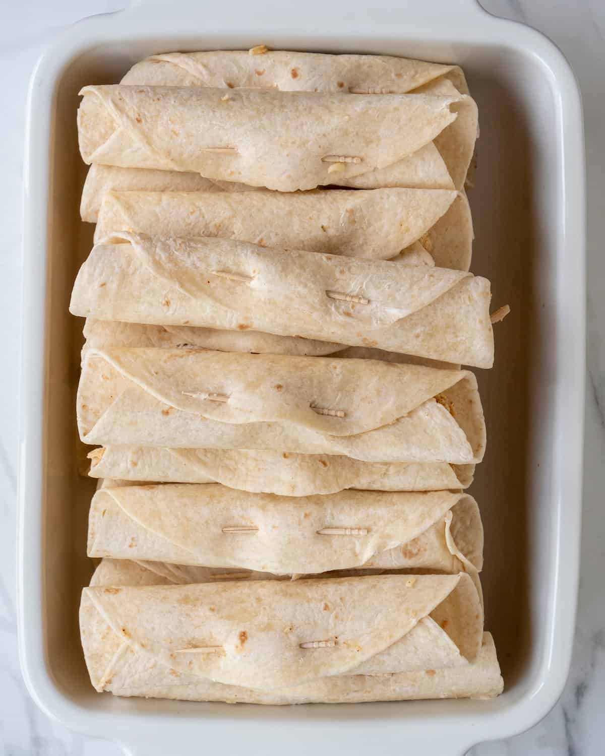 A baking dish with rolled tortillas filled with the chicken mixture, and secured with a toothpick weaved through the overlapping part to seal them, several rolled tortillas placed one on top of the other.