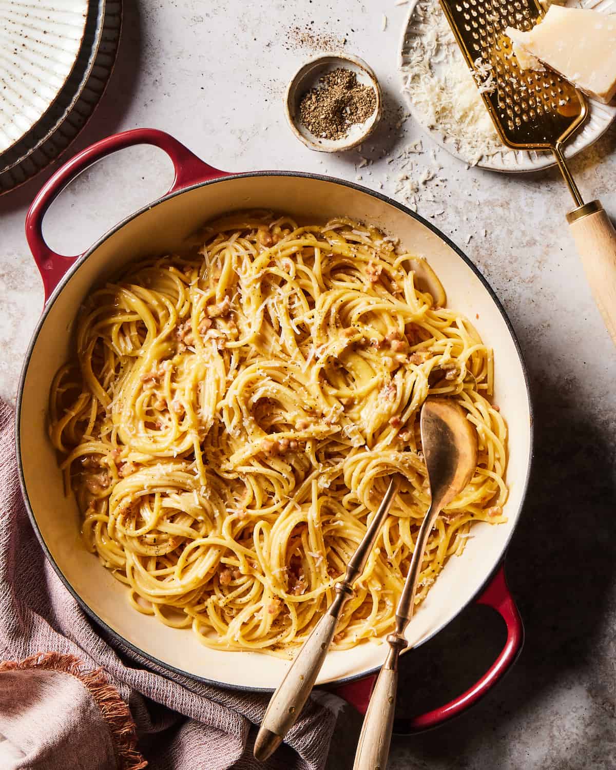 Spaghetti Carbonara in a red dutch oven skillet with two serving spoons beside three stacked ceramic plates in the top left corner and a ceramic bowl of cracked black pepper, and a cheese grater resting on a ceramic plate with grated parmesan cheese in the top right corner.