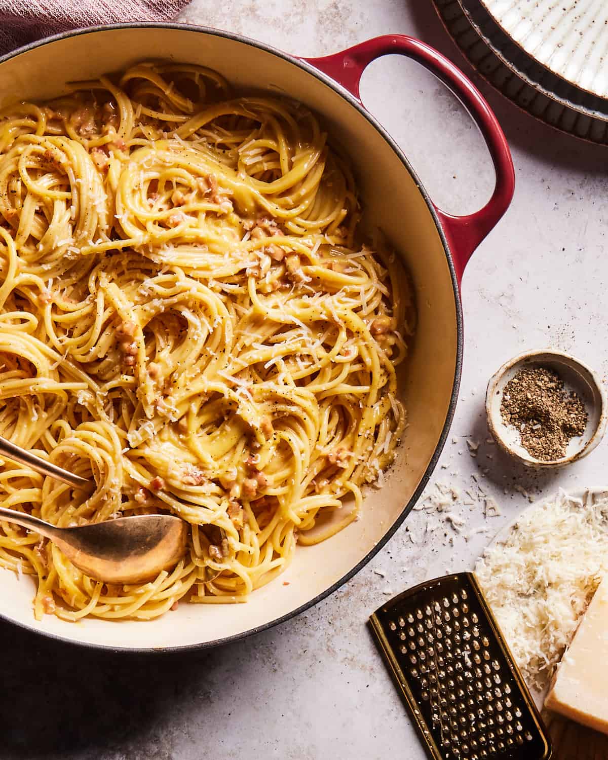 Spaghetti Carbonara in a red dutch oven skillet with two serving spoons beside three stacked ceramic plates in the top right corner and a ceramic bowl of cracked black pepper, and a cheese grater resting on a ceramic plate with grated parmesan cheese in the lower right corner.