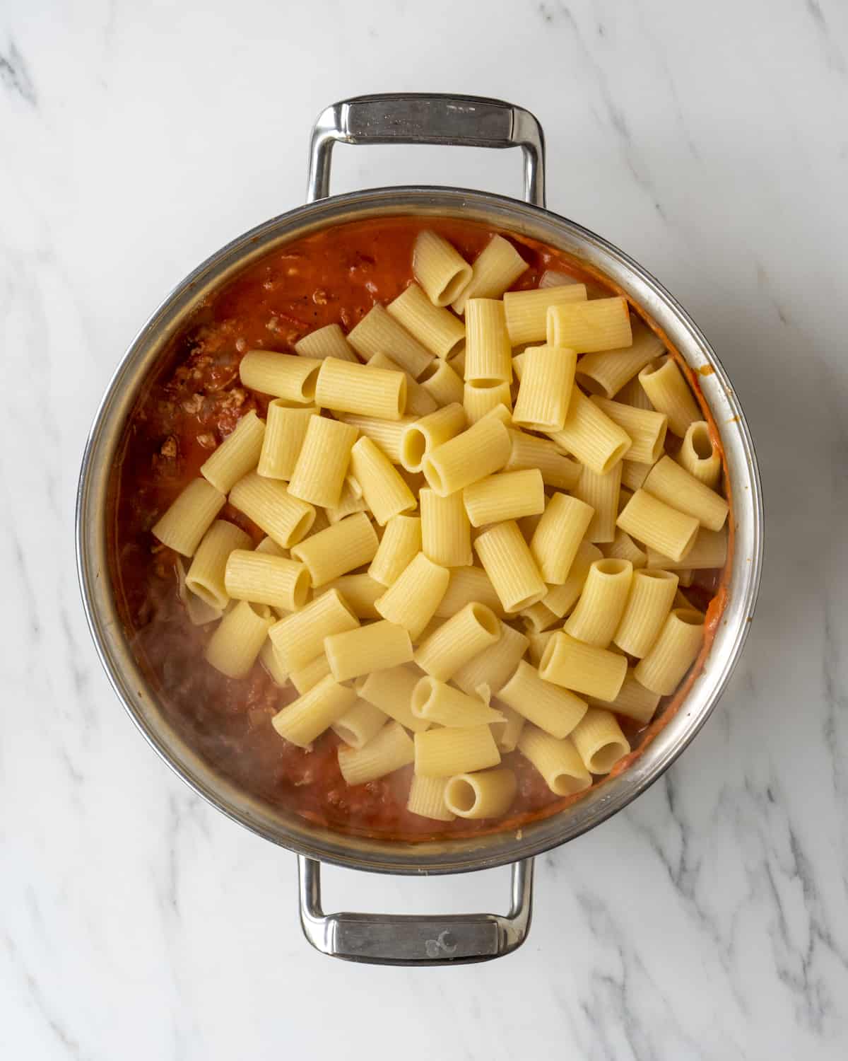 A skillet with red sauce with sausage being cooked and boiled rigatoni pasta just added to the skillet.