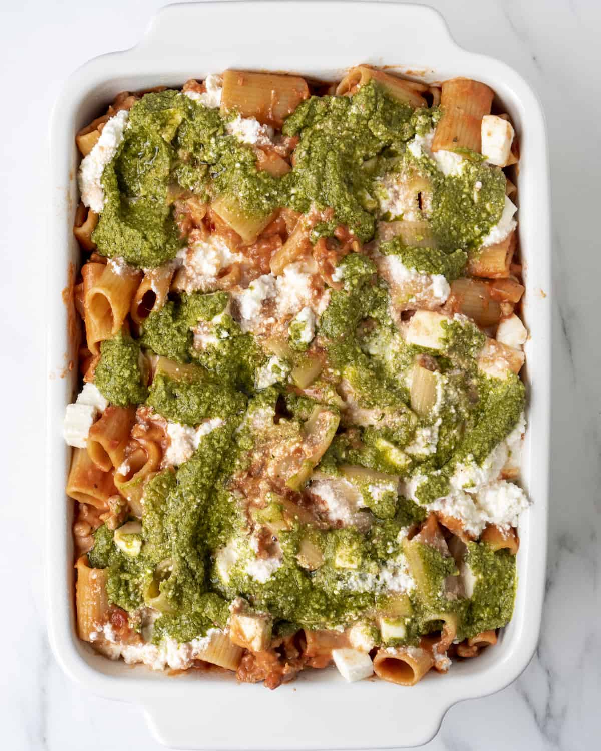 A white rectangular baking dish with rigatoni mixed in red sauce in the bottom, topped with mozzarella, ricotta and pesto.
