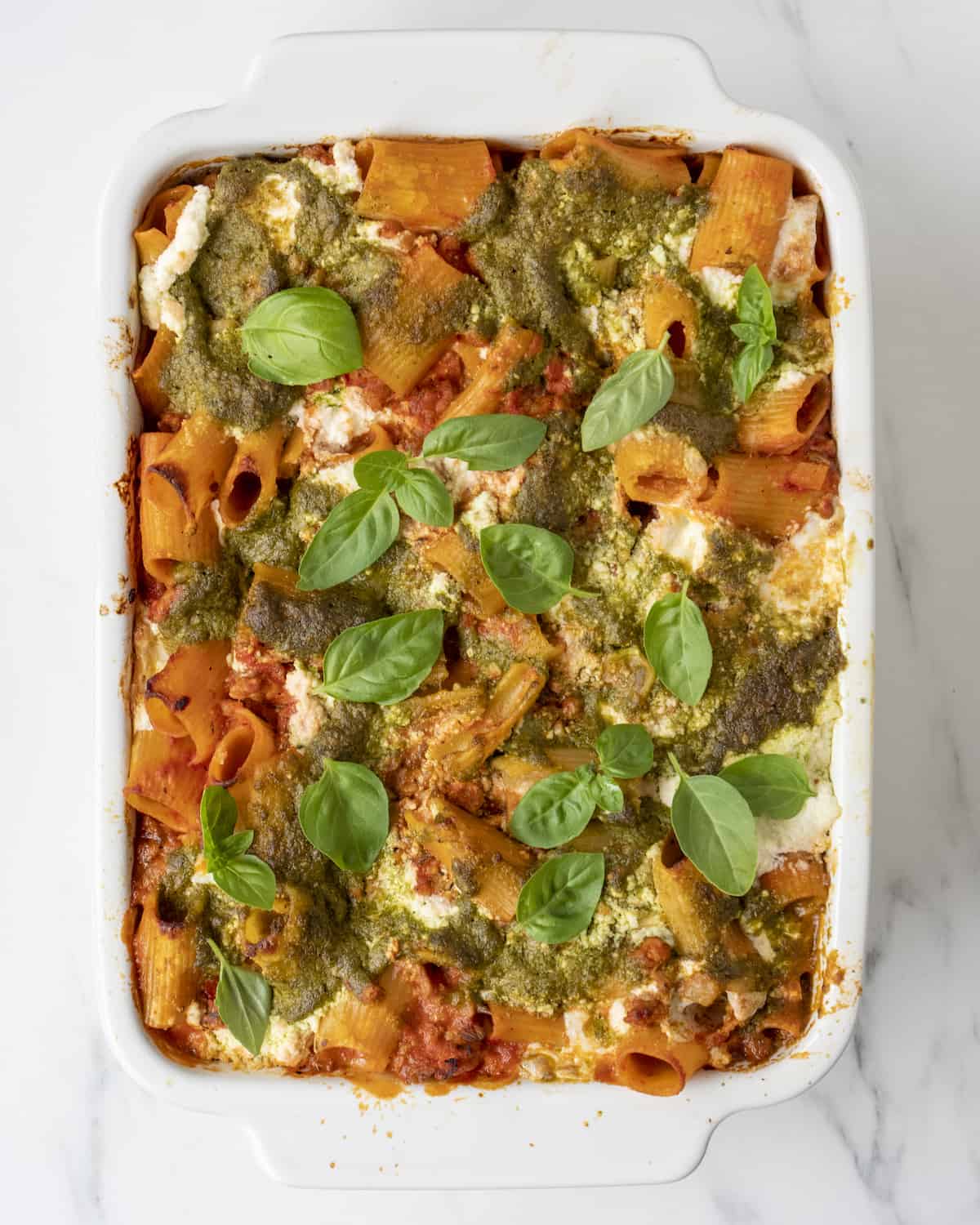 A white rectangular baking dish with baked rigatoni with pesto and ricotta, garnished with basil leaves.