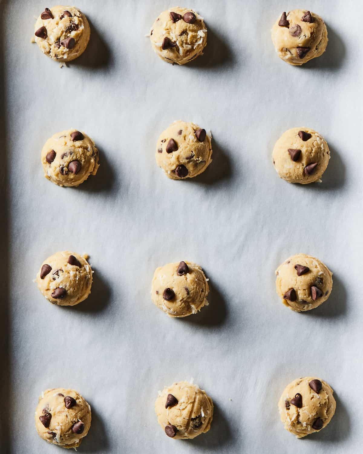 A parchment lined surface with a 4x3 grid of Coconut Chocolate Chip Cookie dough balls.