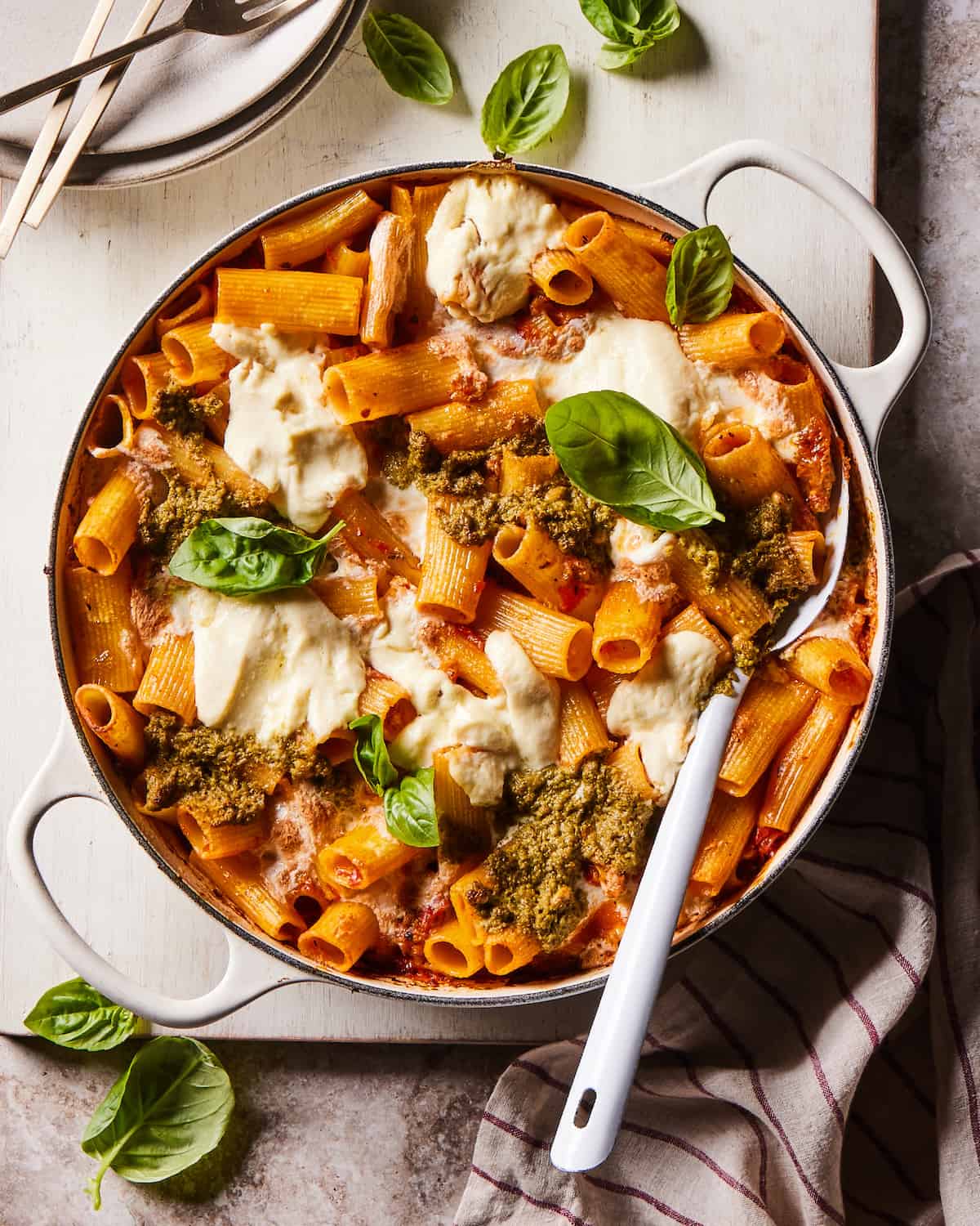 A white skillet with baked Rigatoni with ground chicken sausage, pesto and ricotta garnished with basil leaves, with a serving spoon, and placed on a wooden board and a kitchen towel with more basil leaves on the work surface.