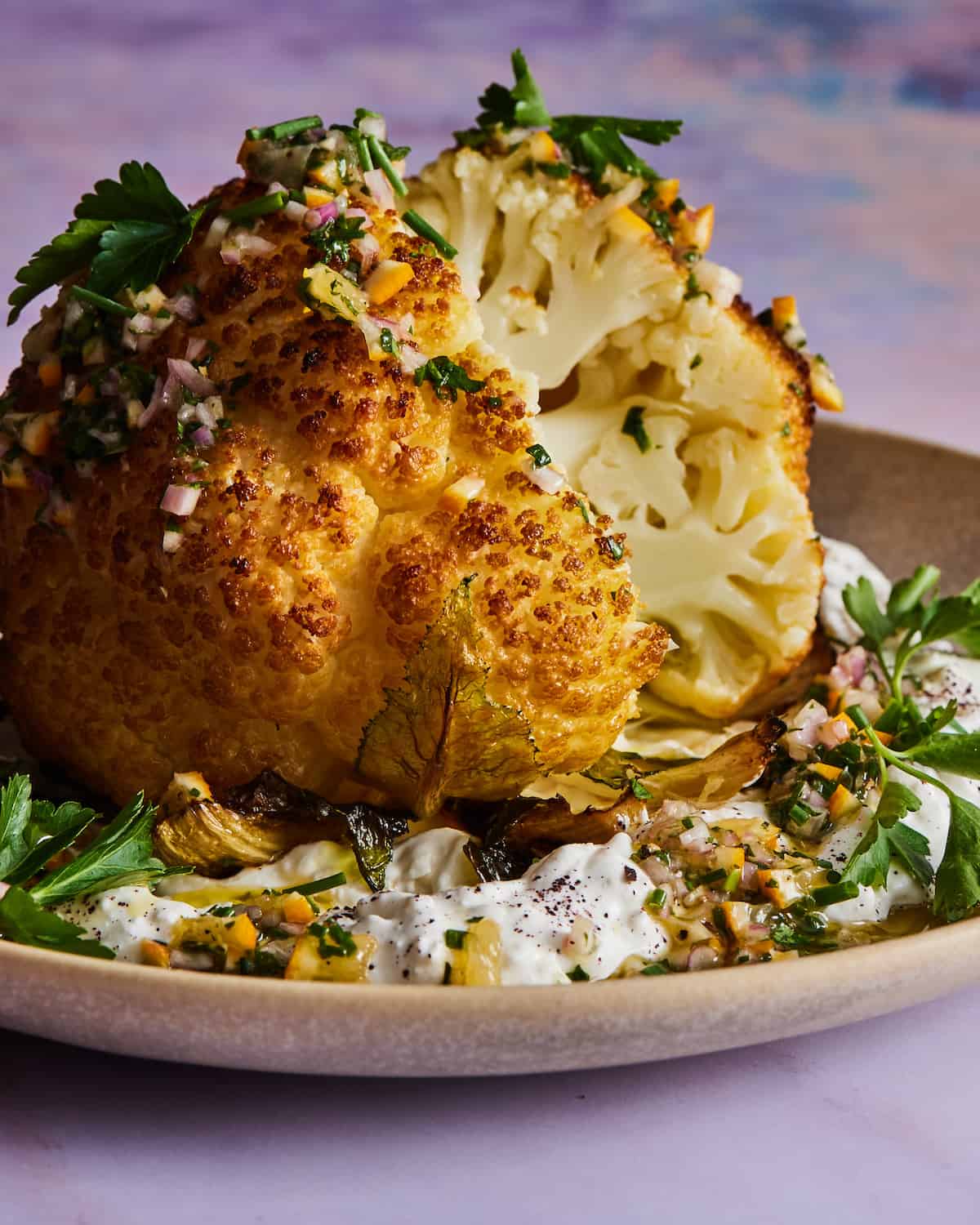 A whole roasted cauliflower garnished with herbs placed on top of a bed of labneh in a beige color ceramic plate.