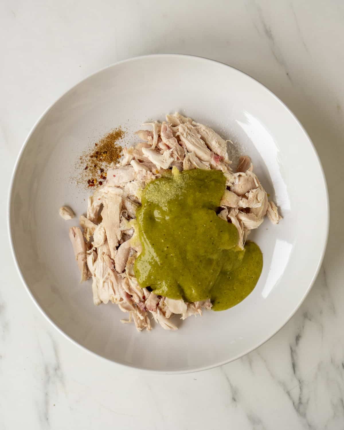Rotisserie chicken ,coriander and red pepper flakes sitting in a white bowl covered with the green enchilada sauce.  
