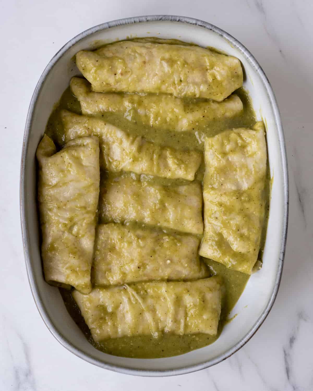 8 enchiladas filled with chicken sitting in a baking dish covered in the green enchilada sauce.  