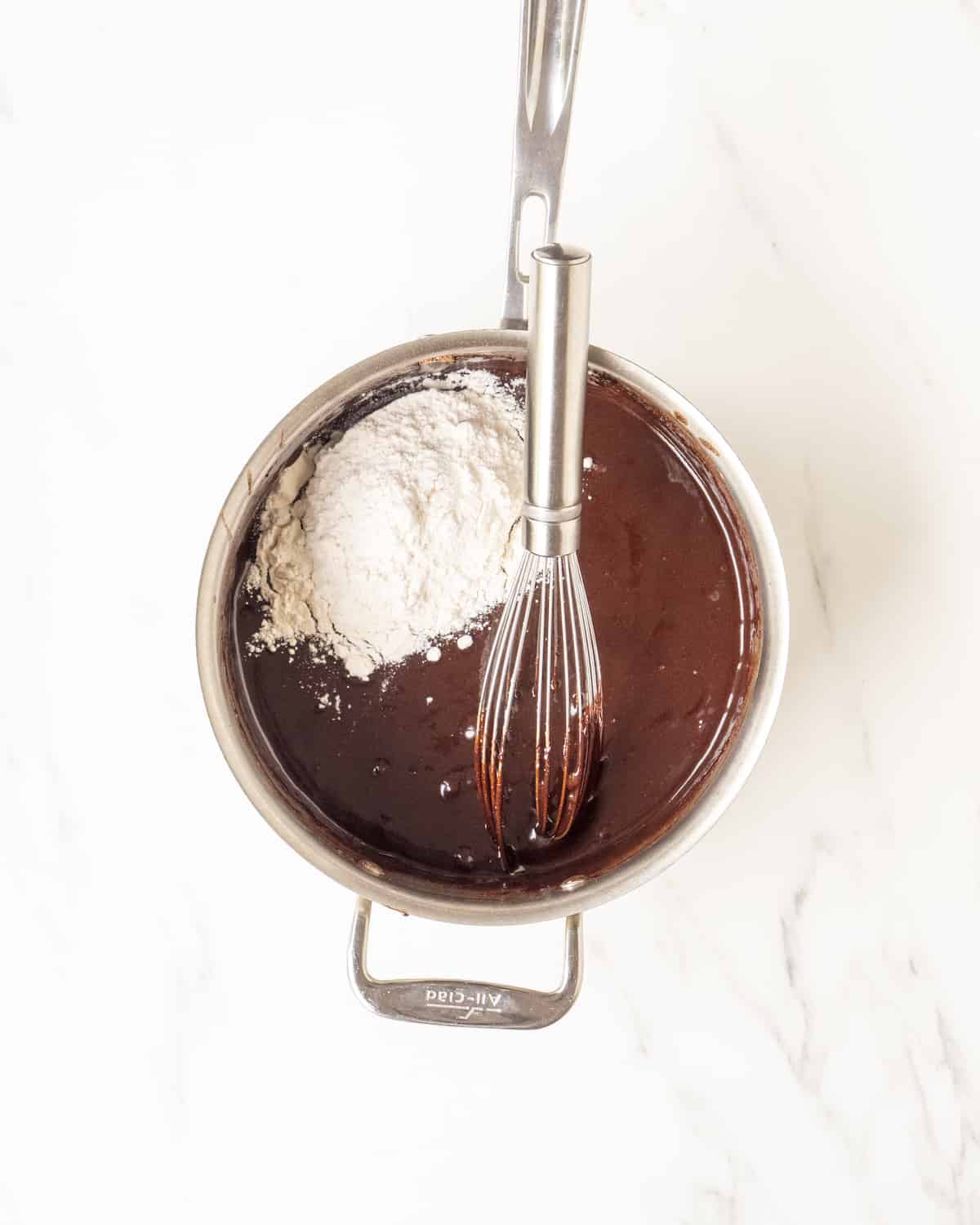 A pot filled with brownie batter, flour, and a whisk.  