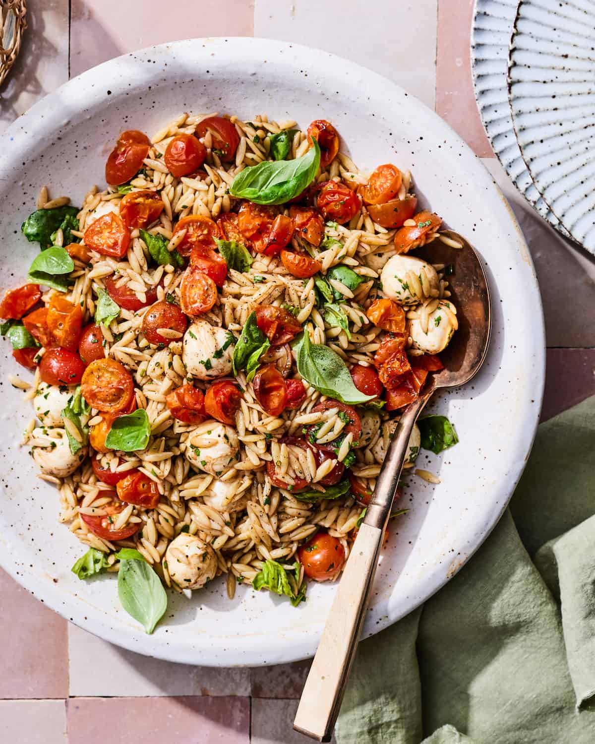 A large gray ceramic bowl of orzo pasta salad with jammy tomatoes, fresh basil leaves, and small mozzarella balls in a balsamic vinaigrette on a pink tiled table with a green napkin in the lower right corner and a stack of two ridged gray ceramic plates in the upper right Corner. 