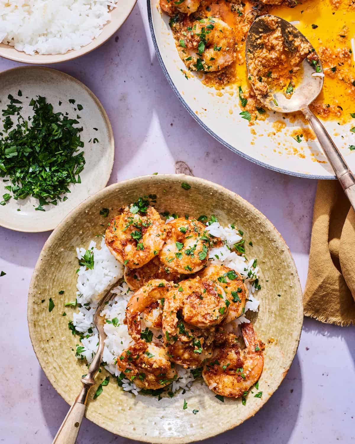 An overhead shot of a bowl of cooked Hawaiian garlic shrimp over white rice with a garnish of minced parsley. There is a fork nestled in the rice on the left hand side of the shallow ceramic bowl. Above the bowl you can see a a small plate of minced parsley, a bowl of white rice, and the remaining garlic shrimp in the enameled cast iron pan ready to be assembled.