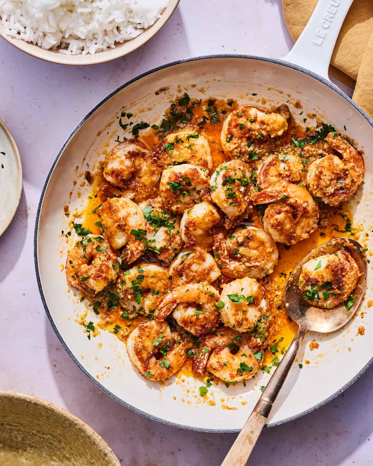 Overhead shot of cooked shrimp in a chili garlic butter sauce with a garnish of minced parsley and a spoon sitting on the right edge of the enameled cast iron skillet, with a small bowl of white rice on the top left corner on the side of the skillet.