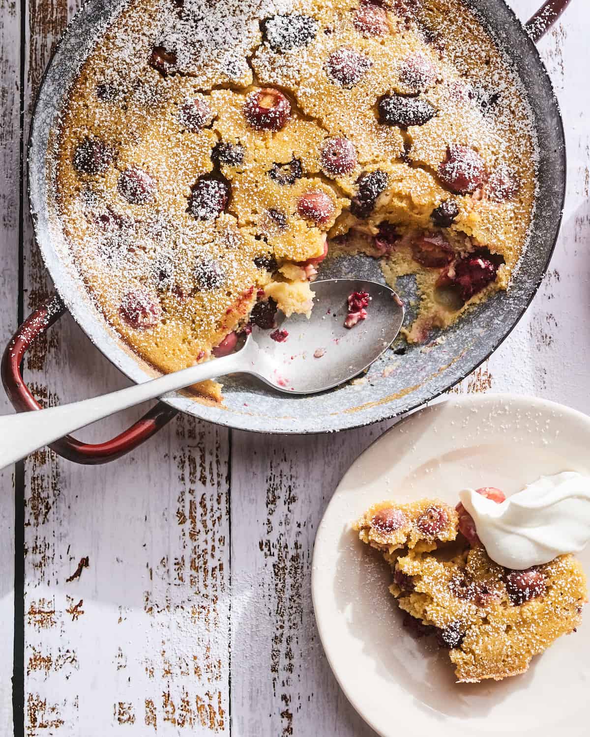 Cherry Clafoutis in a round copper metal baking dish on a white washed wood background with a dusting of powdered sugar sprinkled over top and a spoonful or two missing from the bottom right corner of the baking dish and a serving of cherry clafoutis on a plate in the bottom right corner with a healthy dollop of whipped cream.