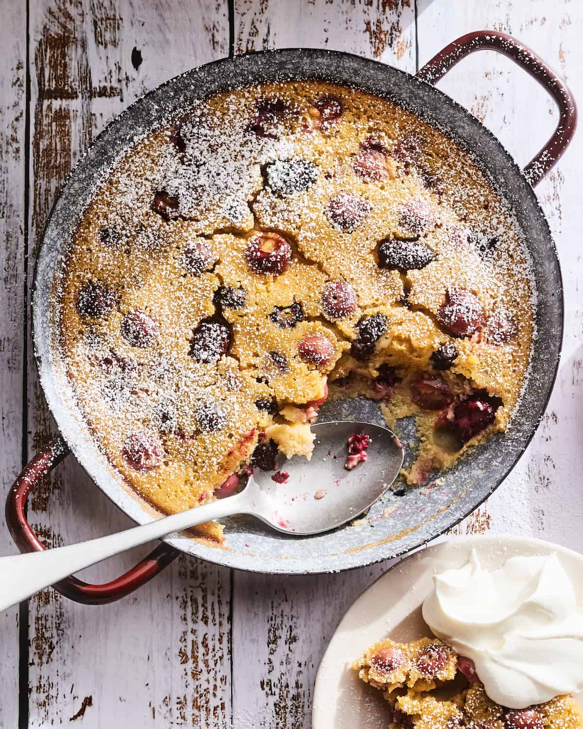 Cherry Clafoutis in a round copper metal baking dish on a white washed wood background with a dusting of powdered sugar sprinkled over top and a spoonful or two missing from the bottom right corner of the baking dish.