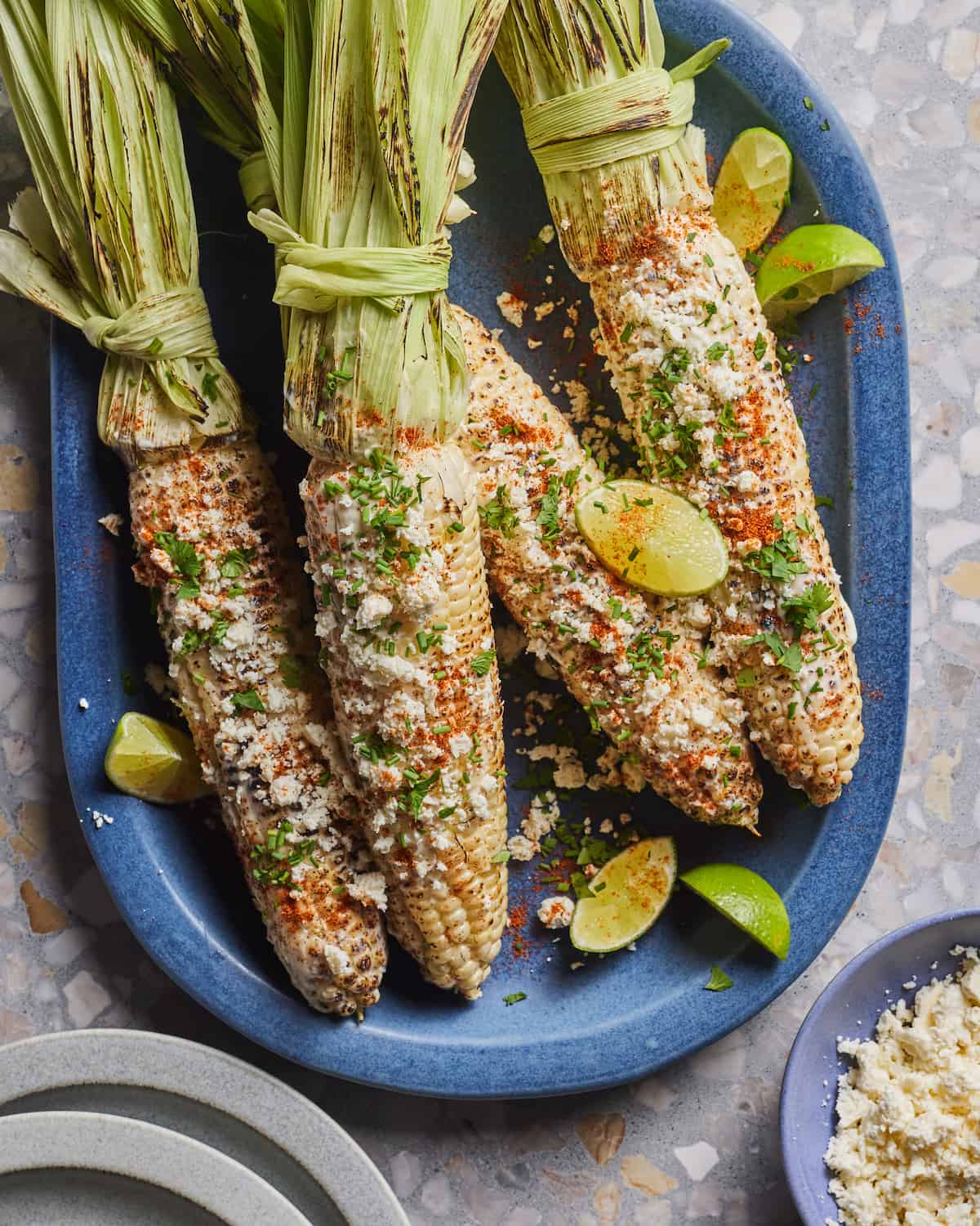 4 grilled cobs of corn covered in feta, cilantro and chili mixture placed on a blue oval platter, with lime wedges, with a small bowl of feta and a couple small plates on the side.