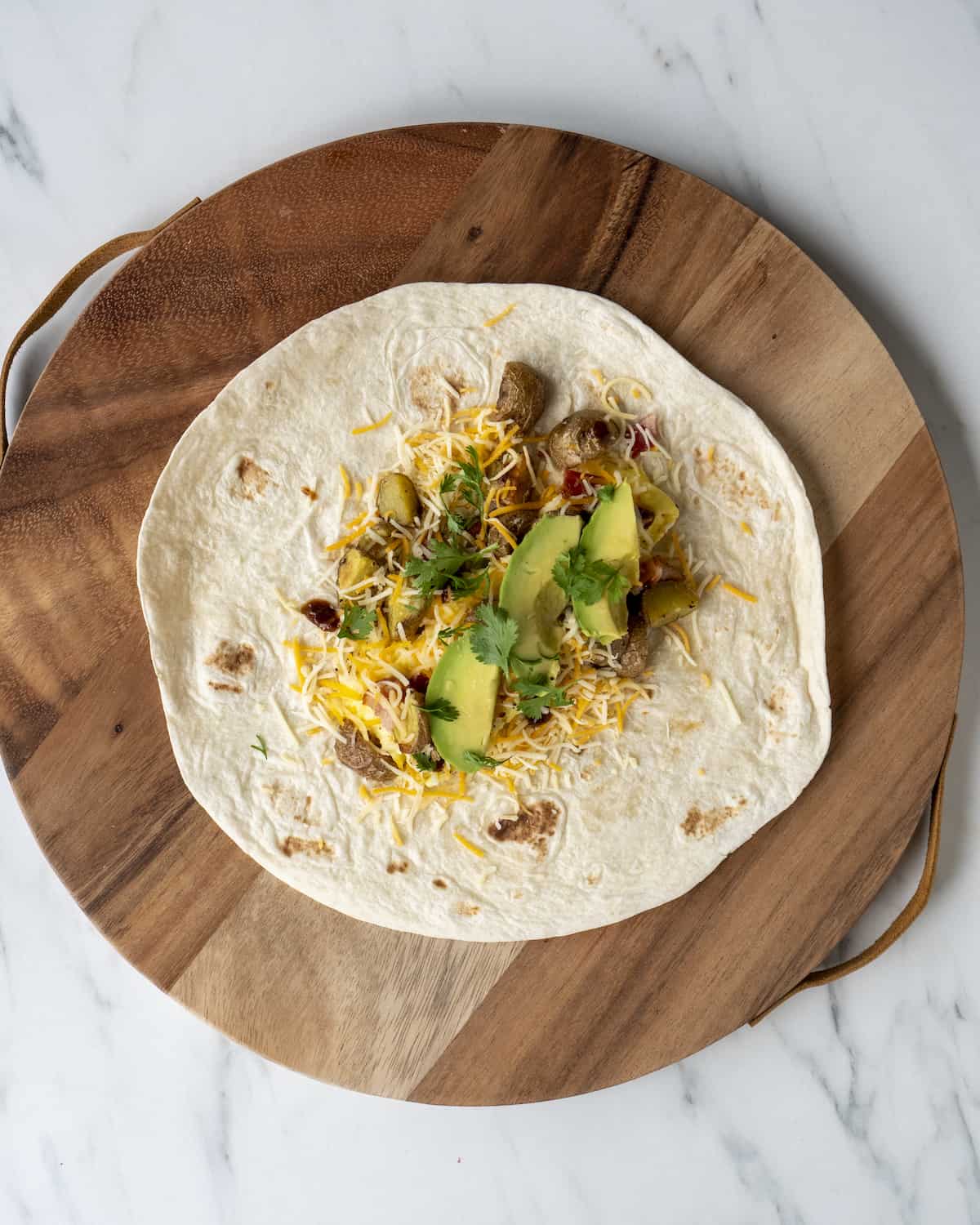 A flour tortilla with the the scrambled eggs, avocado, cheese, roasted potatoes, and cilantro sitting on a wooden cutting board.  
