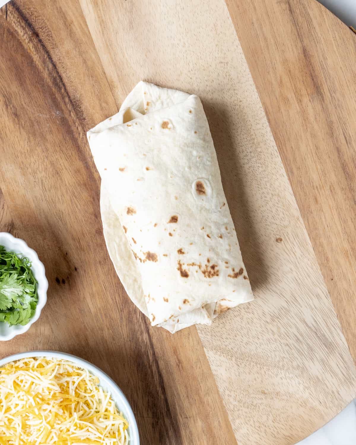 A rolled up breakfast burrito sitting on a wooden cutting board next to a bowl of shredded cheese and cilantro.  