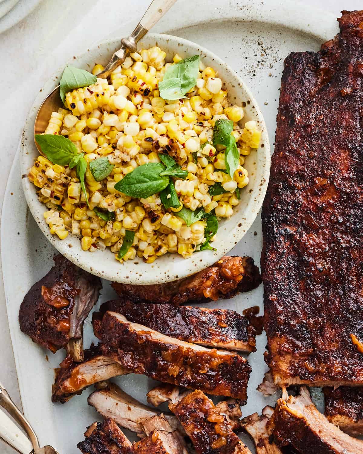A bowl of charred corn with leaves of basil sitting next to a rack of BBQ ribs. Makes the perfect BBQ Corn Salad