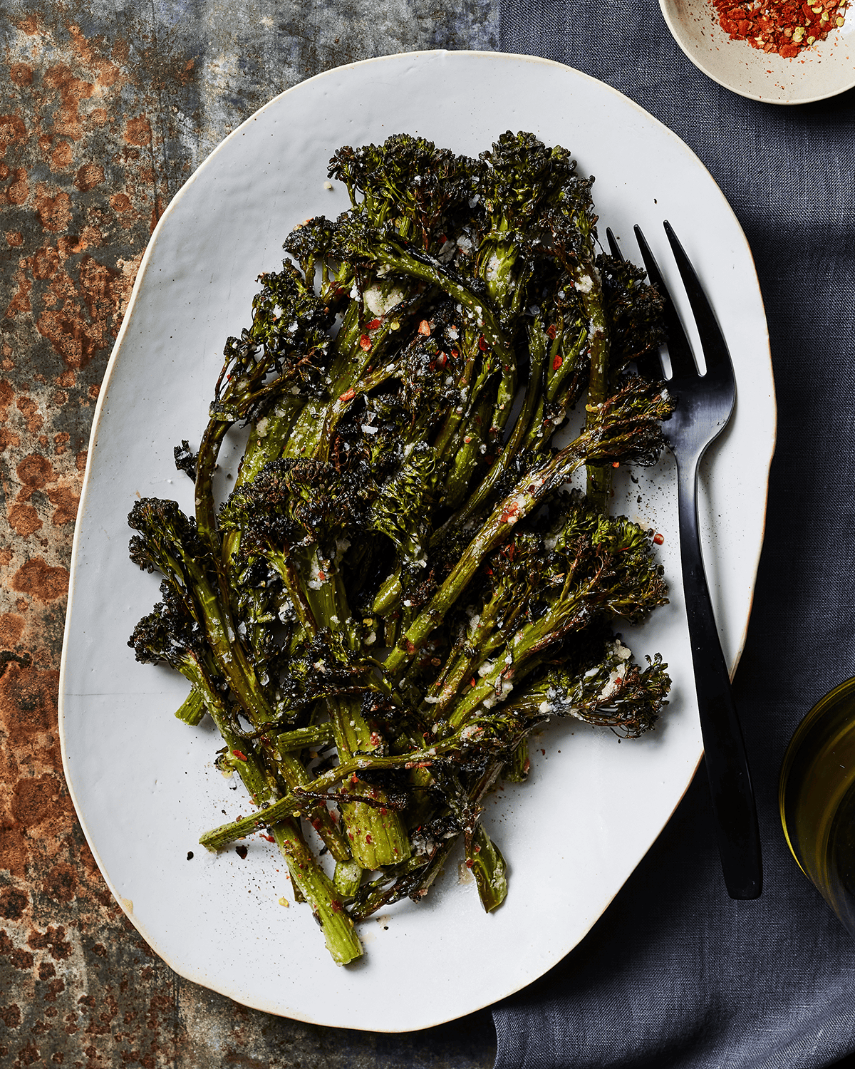 Roasted Broccolini with red pepper flakes sitting in a bowl with a fork next to a bowl of red pepper flakes on a table top.  