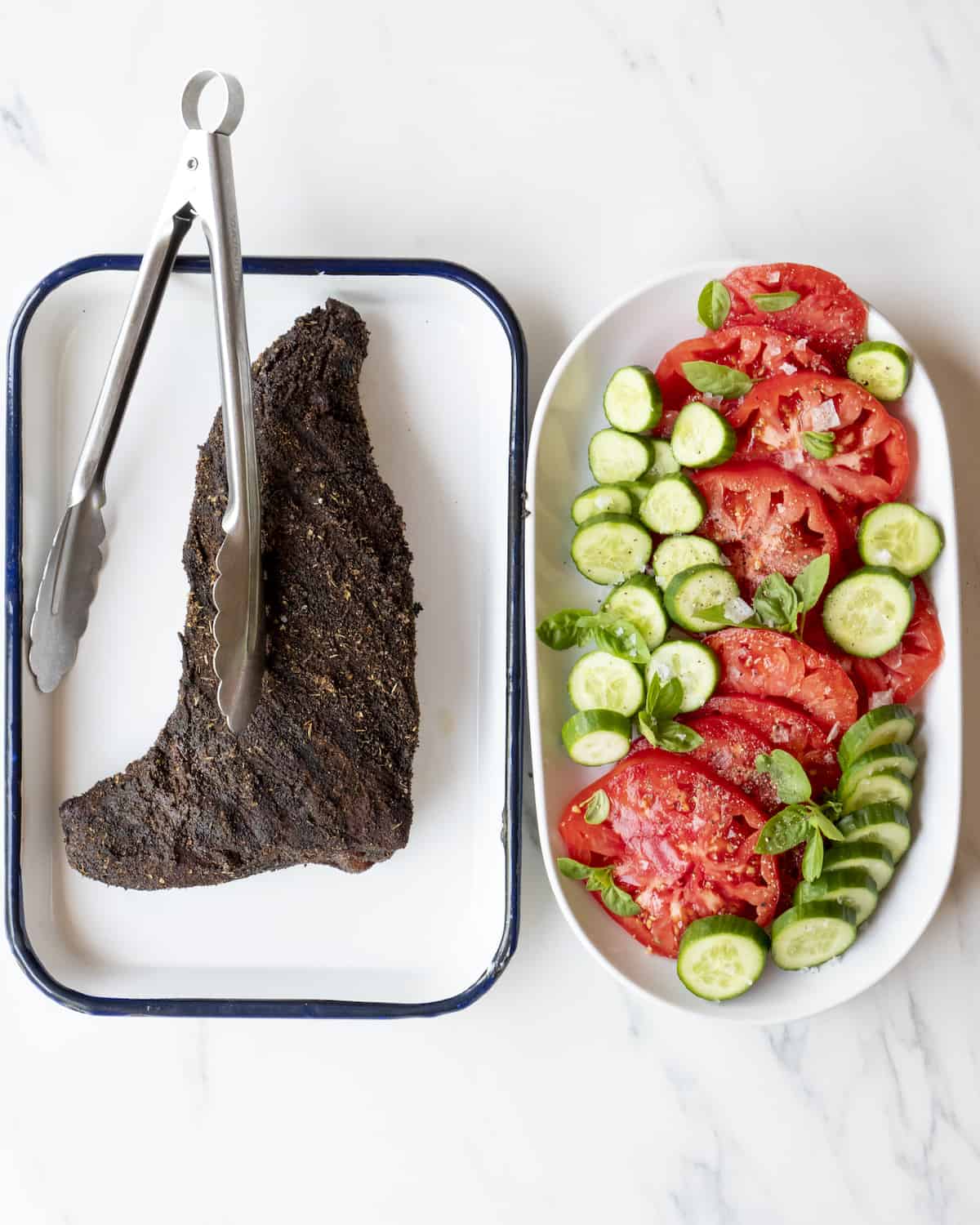A trip tip that has been seared is sitting in a baking dish next to a dish full of tomatoes basil, and cucumbers.  