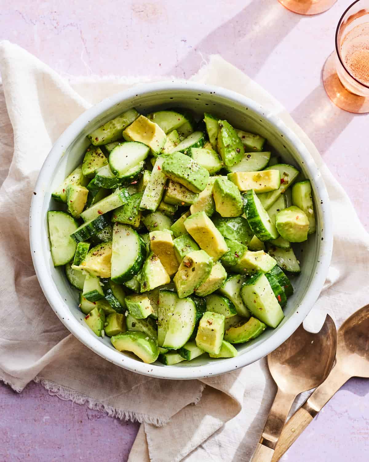 Cucumber salad in a ceramic bowl with sliced cucumbers and avocados mixed together in a vinaigrette. The bowl is on a cream linen napkin on a lavender table with two gold serving spoons in the bottom right corner and two orange glasses in the the top right corner