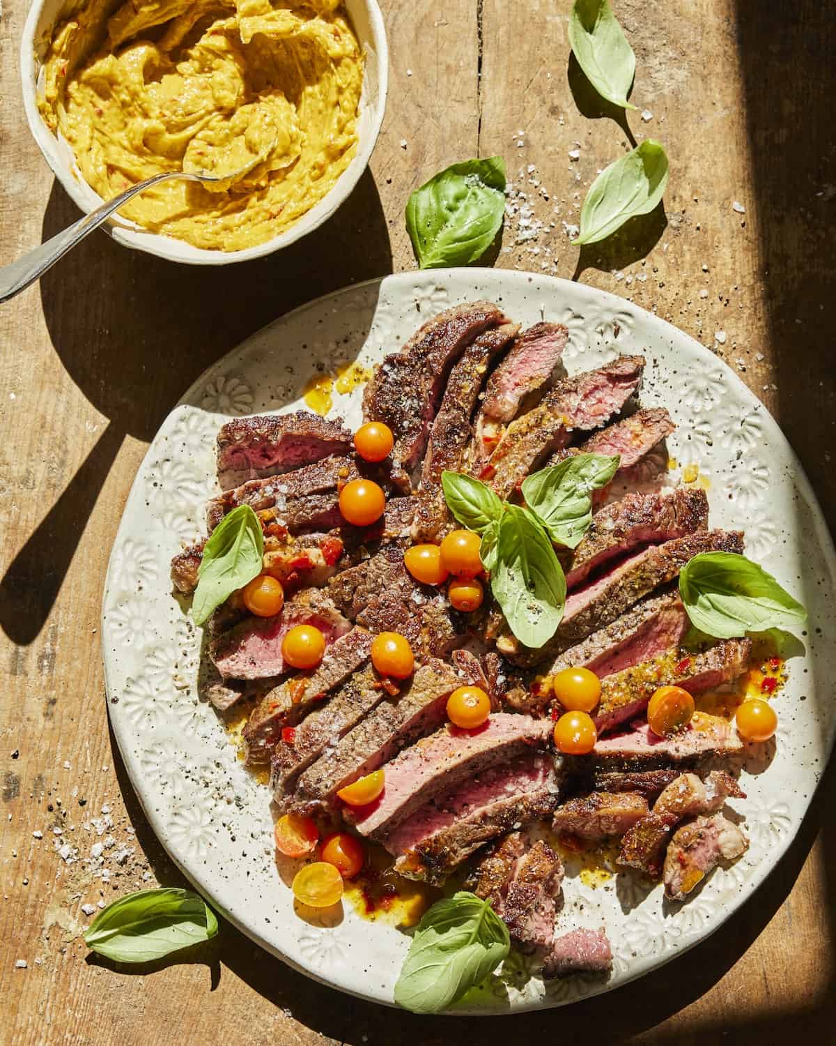 Strips of cut ribeye sitting on a plate topped with halved cherry tomatoes and leaves of basil.  The plate is sitting next to a dish of Calabrian chile butter.