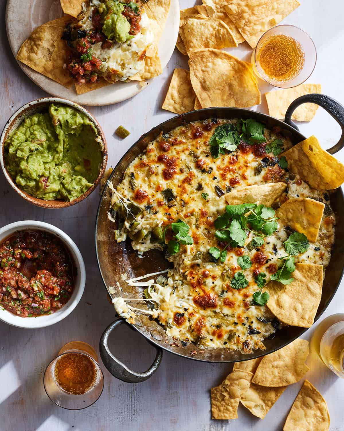 A tabletop with a skillet of queso next to bowls of guacamole, salsa, a beer, and a plate full of nachos.  