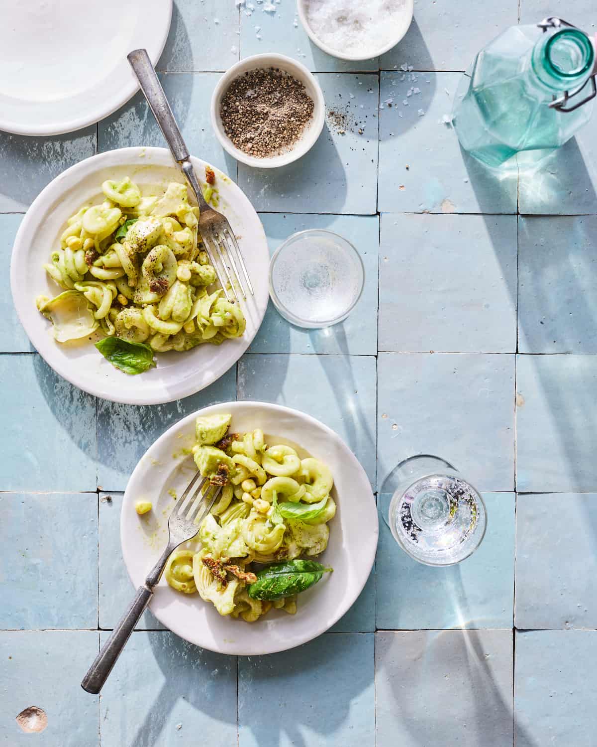 Two plates of artichoke pasta sitting next to each other on a blue tile table.  