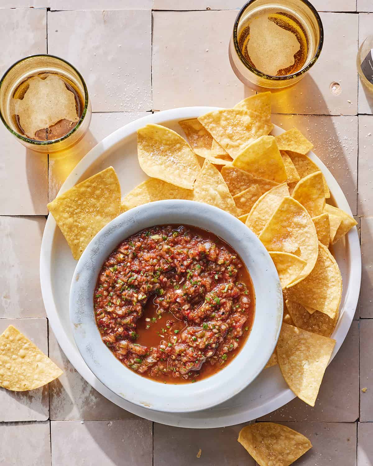 A bowl of red salsa sitting on a plate next to some tortilla chips and pints of beer.  